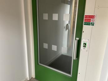 Mobility lift door with green surrounding wood and vision panel with clear manifestations at dual height.
