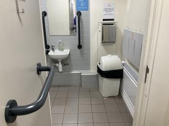 Accessible toilet showing outward opening, white door with contrasting blue grab rail , waste bin and sink.