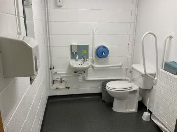 View of accessible toilet facility showing toilet with right hand transfer and adjacent grab rail, plus sink and hand dryer.