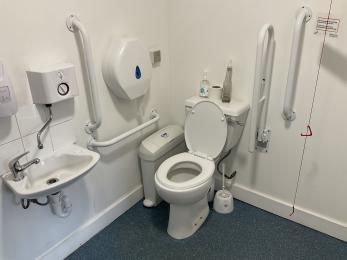 White toilet showing right hand transfer, sink and white grab rails against white walls.