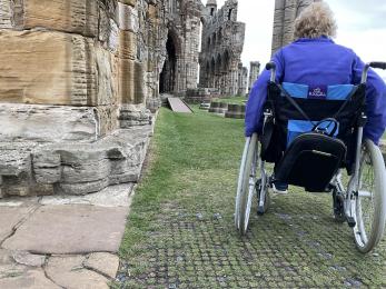 Wheelchair user on matting surface over grass in Abbey grounds with wooden access ramp and Boardwalk in the background.