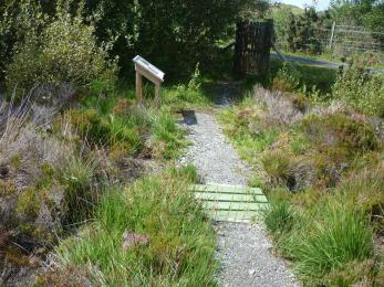 A narrow, gravel path winds through vegetation. There's a small bridge which is on a level with the rest of the path. 
