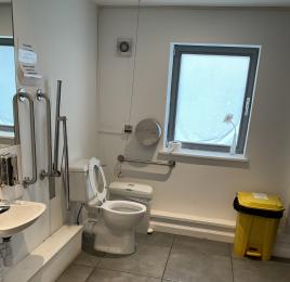 Accessible white toilet with white sink and silver grab rails and large floor space.