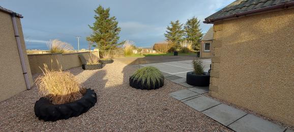 Image is of pebbled area with tractor tyre planters with the carpark & lawned area beyond