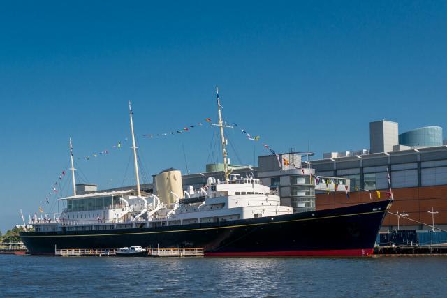 An exterior side view of The Royal Yacht Britannia berthed at Leith's Ocean Terminal against a bright blue sky. 