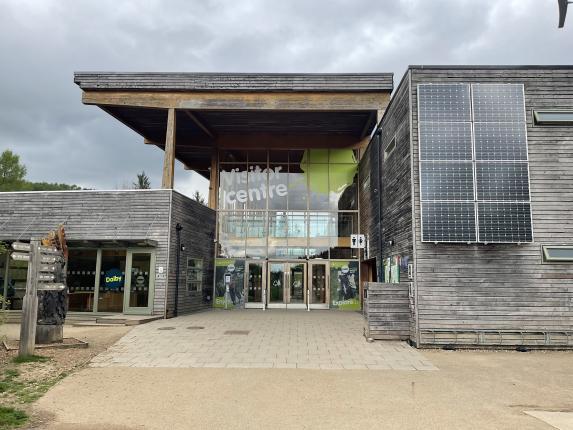 Wide pathway leading from car park into Dalby Forest  visitor centre, with glazed doors and window panels, surrounded by forest.
