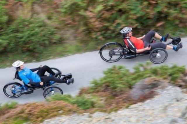 Two people riding adapted cycles through forest trails