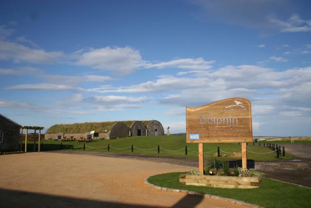 The image shows the Scottish Dolphin Centre visitor centre, including historical buildings, a grassy hill and blue skies